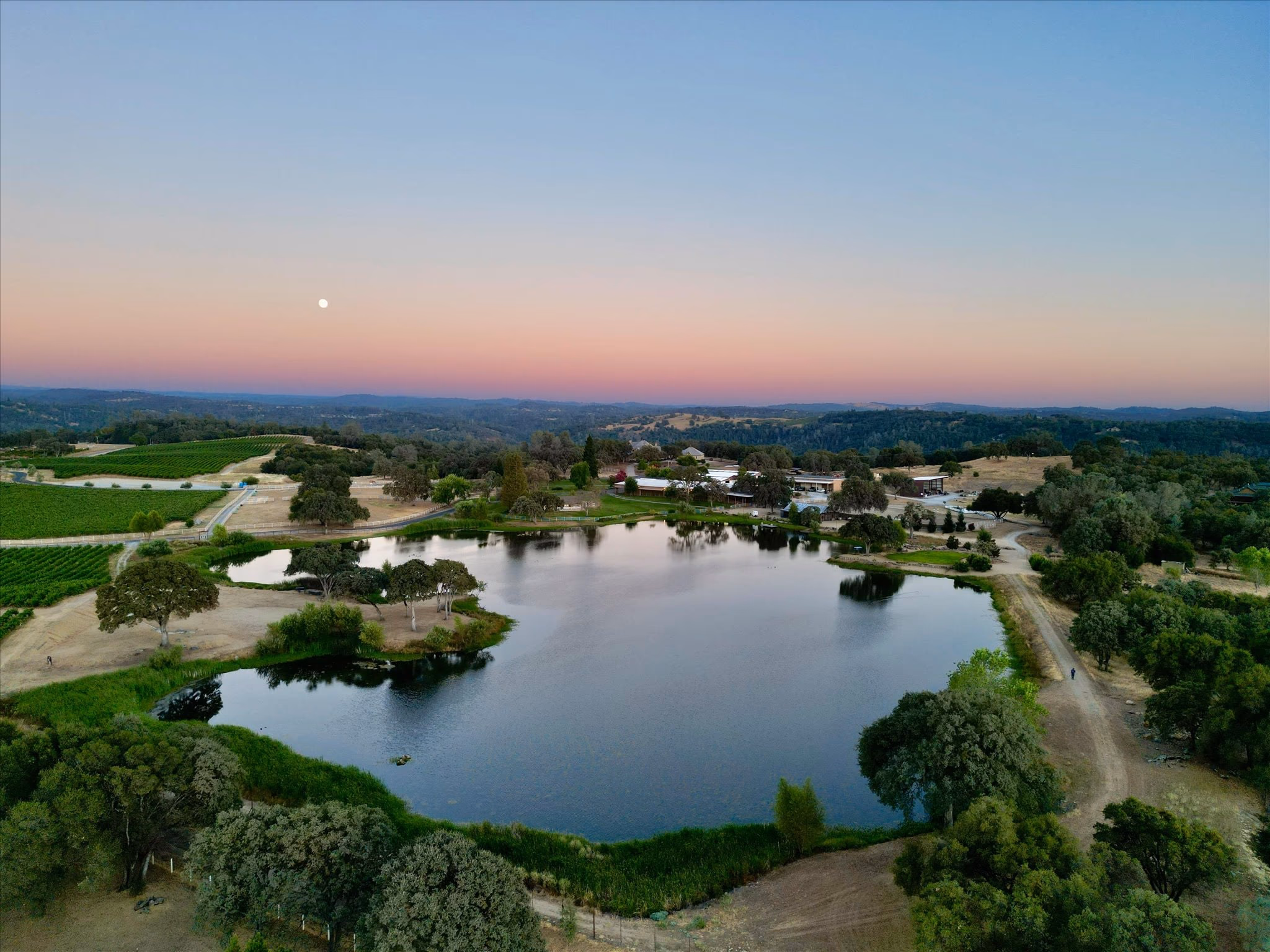 The private nine-acre lake at Saureel Vineyards at dusk, seen from the air, with vineyard blocks, the ranch buildings and the Sierra foothills beyond.