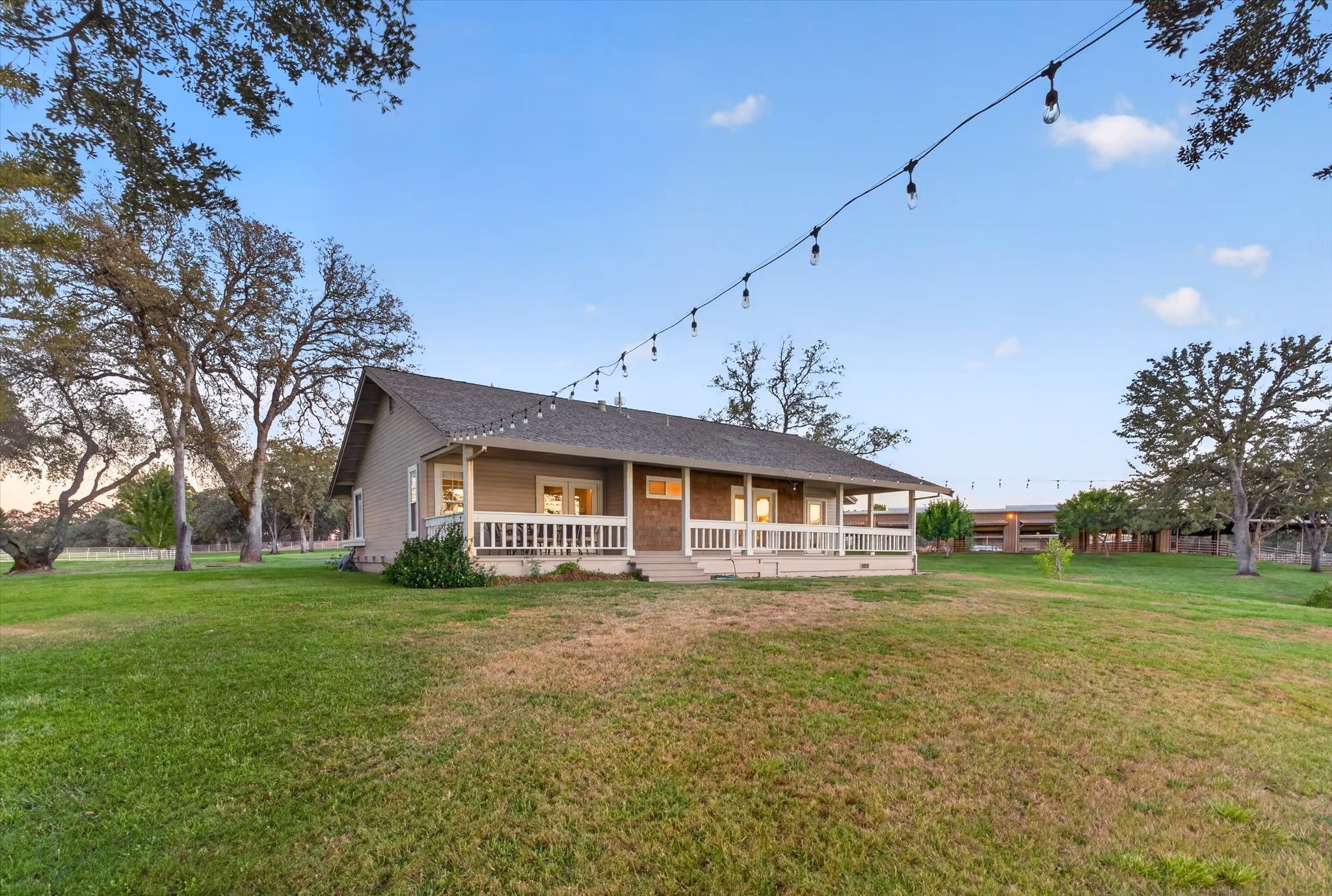 The lake house, a single-story house with a covered porch, seen across a broad lawn with festoon lighting strung between oaks.