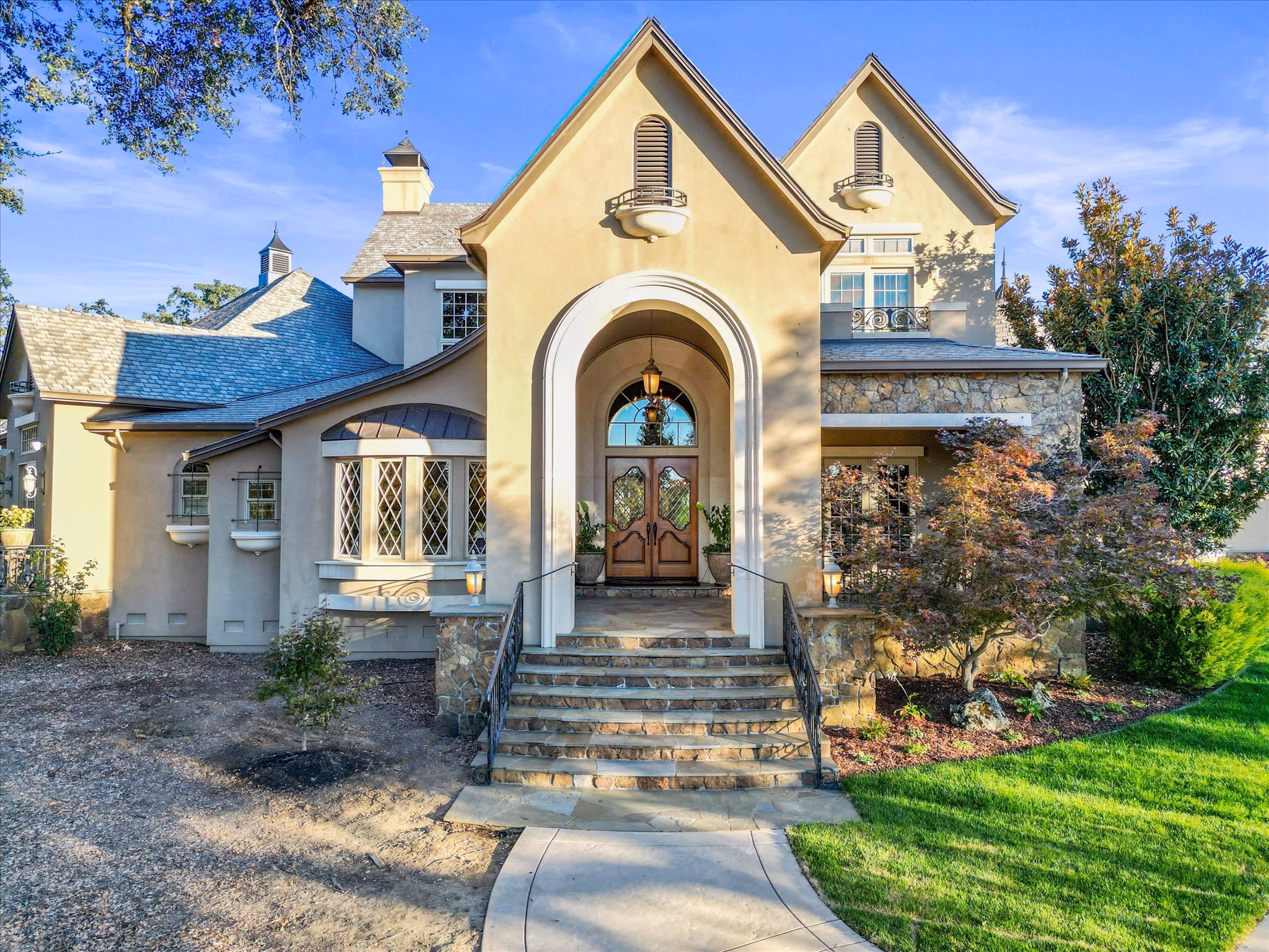 The arched stone entry of the main residence in daylight, with leaded bay windows, a slate roof and stone steps up to double timber doors.